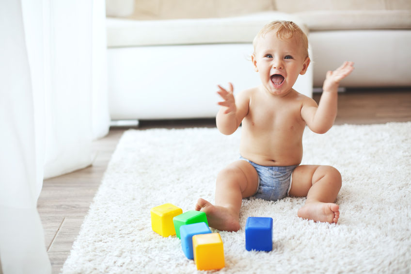 An image of a happy baby sat on a rug wearing a nappy and looking excited